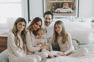 Family wearing matching beige and white gingham check pajama sets sitting on a bed.