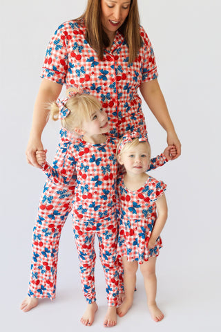 Woman and two children wearing matching pajama sets with cherry and gingham print in red, white, and blue.