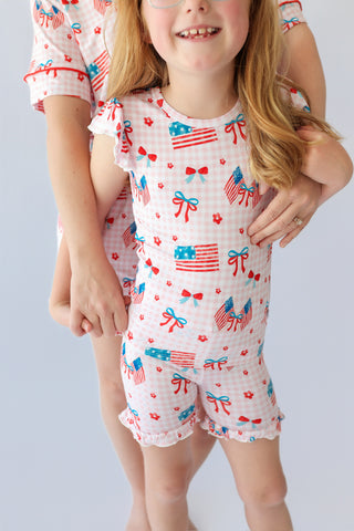 Child wearing white short set with red, blue and white flag, bow, and flower print throughout with ruffled sleeves and shorts