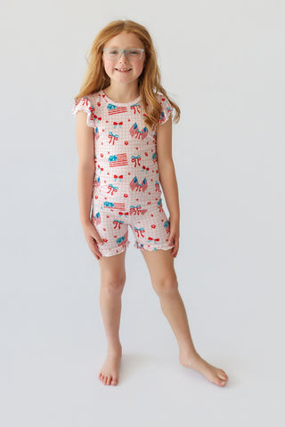 Girl wearing short set with ruffled sleeves and legs, featuring red and blue flag and bow print on white background.