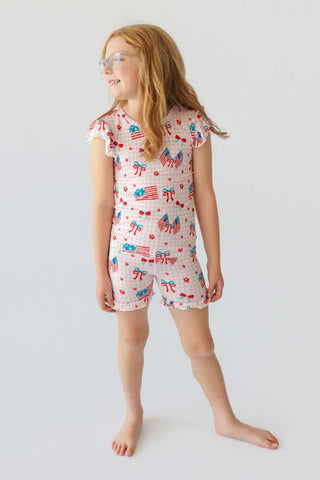 Child wearing light pink short set with red bows, flags, and cherries print, ruffled cap sleeves and shorts.