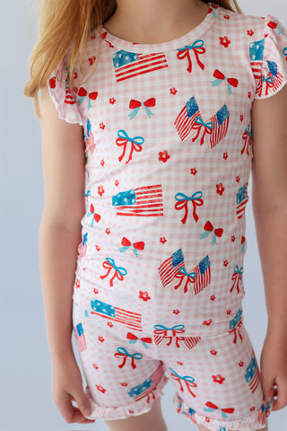 Pink gingham short set with American flag, bow, and flower print, ruffled neckline and sleeve cuffs.