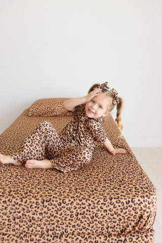 Child wearing leopard print pajamas sitting on matching leopard print twin sheet set with pillow.