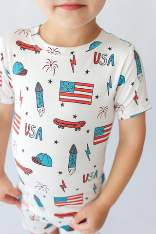 Child's cream short-sleeve t-shirt with red, white, and blue skateboard, flag, rocket, and fireworks print.