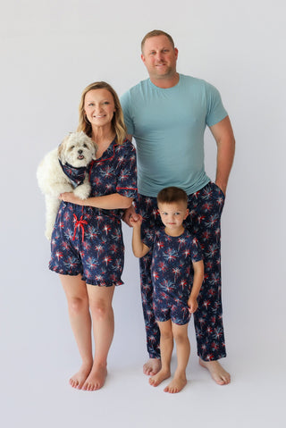 Woman wearing navy shorts with red and white firework pattern, holding white dog, standing with man and child in matching paj