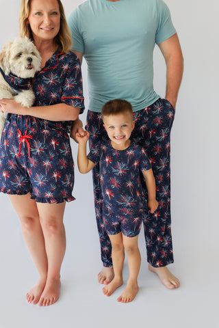 Woman and child wearing navy shorts with red and white firework print; man in blue shirt and matching shorts; small dog in wo