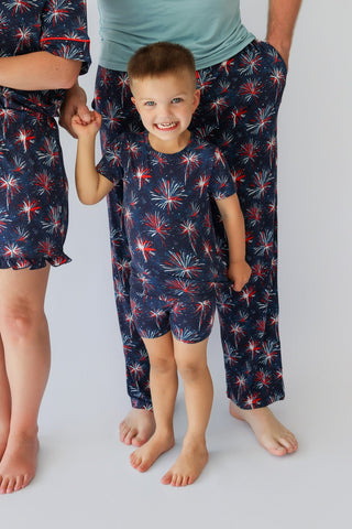 Child wearing navy shorts set with red and white firework pattern, standing between two adults in matching pajamas.