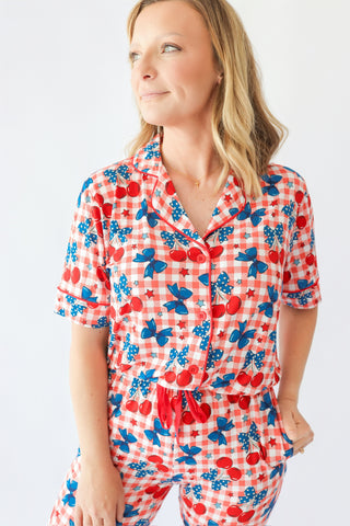 Woman wearing short-sleeve pajama top with red and white gingham, cherry, butterfly, and star print pattern with button front