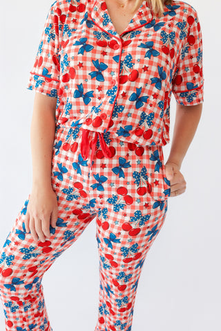 Woman wearing red and white gingham pajama set with cherry and blue bow print, featuring button-up shirt and matching pants.