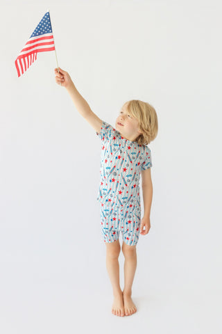 Child wearing light blue short set with red stars and rocket print, holding American flag.