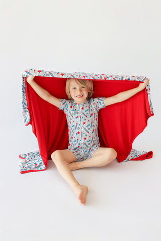 Child sitting under red blanket with rocket and star patterned border and matching pajamas.