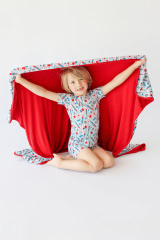 Child holding a red blanket with rocket and star patterned trim on edges.