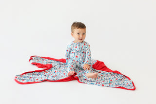 Toddler sitting on light blue blanket with red rocket and star pattern and red binding edges.