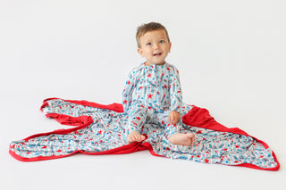Toddler wearing blue pajamas sits on a blue and red rocket-patterned blanket with red trim.