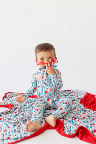 Toddler sitting on blue, red and white rocket-patterned blanket with red trim, wearing matching pajamas.