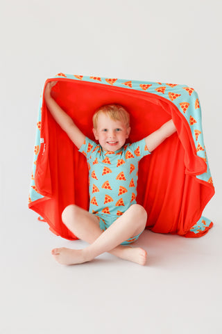 Toddler wearing blue short set with orange pizza slice pattern and matching red blanket.