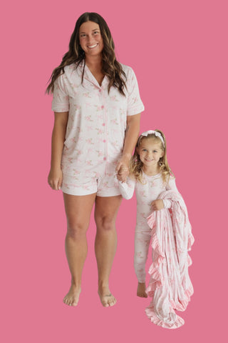 Woman and child wearing matching white short sets with pink floral print and button-up shirts against pink background.