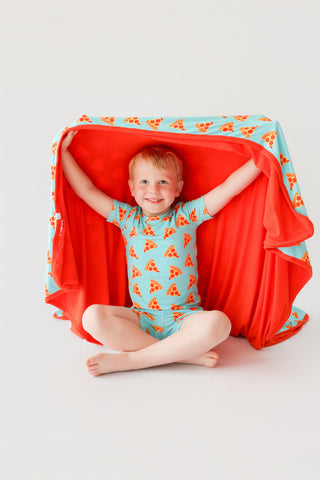 Toddler wearing light blue short set with orange pizza slice pattern and matching red blanket draped overhead.