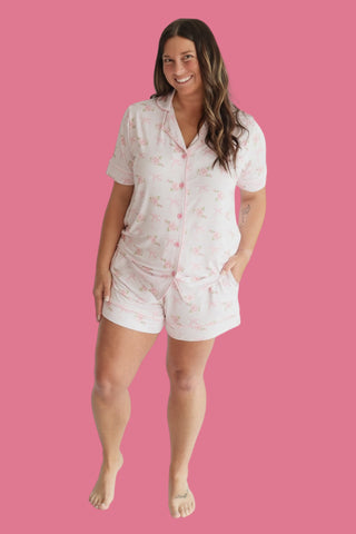 Woman wearing white short-sleeve button-up top and matching shorts with pink floral pattern against pink background.