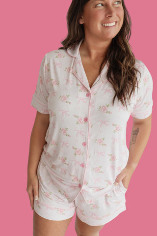 Woman wearing white short pajama set with pink floral and bow print, button-front shirt and matching shorts against pink back