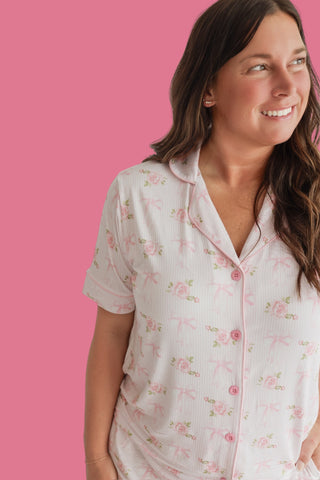Woman wearing white short-sleeved pajama top with pink floral and bow pattern and button front against pink background.