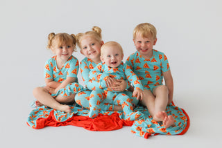 Four toddlers wearing turquoise rompers with red and orange pie slice print and matching red ruffle trim.