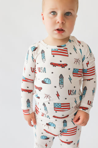 Toddler wearing cream-colored long-sleeve pajamas with all-over print of skateboards, rockets, flags, stars, and "USA" text i