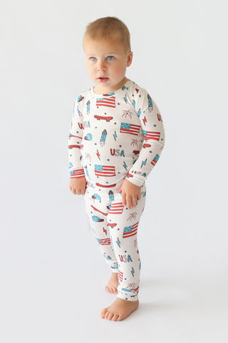 Toddler wearing cream pajamas with American flags, skateboards, rockets, and "USA" text in red and blue.