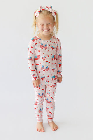 Young child wearing light pink pajama set with red, white, and blue flag and bow print, paired with matching hair bows.