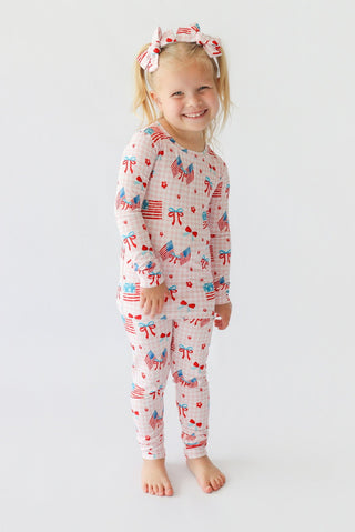 Young child wearing red, white, and blue patterned pajama set with matching hair bows featuring flags, bows, and hearts.