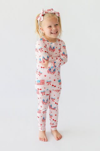 Young child wearing two-piece pajama set with red, white, and blue flag and bow print, with matching hair bows.