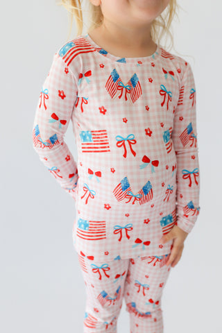 Child wearing pink and white gingham pajamas with red, blue, and white patriotic print including flags, bows, and flowers.