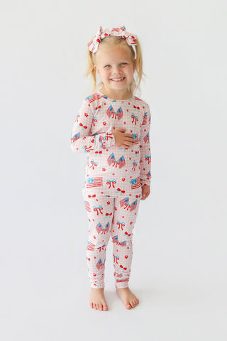 Young child wearing long-sleeved pajama set with flags, bows, and hearts print, paired with matching hair bows.