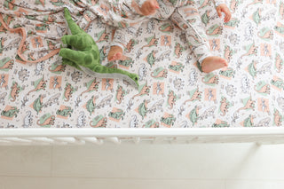 Crib sheet with dinosaur and plant illustrations in muted green, peach, and cream tones, topped with green dinosaur plush toy