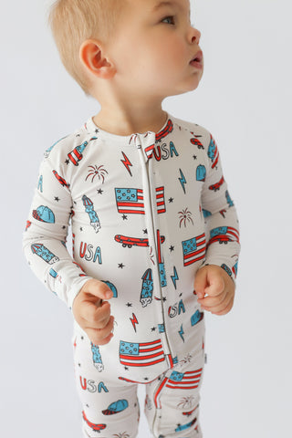 Toddler wearing cream romper with red, white, and blue printed rockets, flags, stars, and "USA" text throughout.