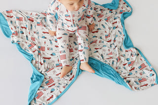 Infant romper with cream base featuring red and blue flags, airplanes, and "USA" text, turquoise trim and lining.
