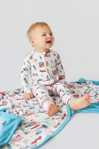 Infant in cream romper with red, white, and blue skateboard and USA flag print, long sleeves, front zip closure.