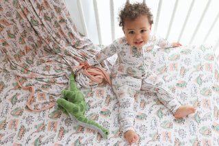 Toddler lying on cream crib sheet with colorful dinosaur and nature print pattern in peach, green, and gray tones.