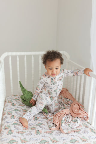 Cream crib sheet with multicolored dinosaur and fossil print in a white crib with toddler.