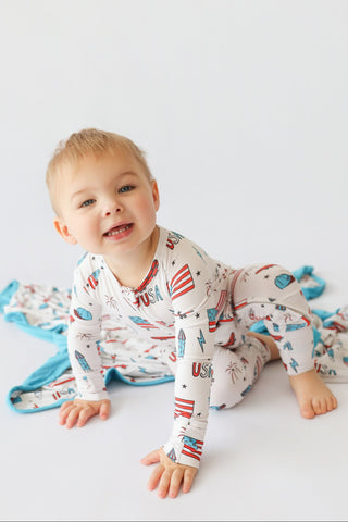 Toddler wearing cream romper with red, white and blue patriotic prints including flags, stars, and surfboards.