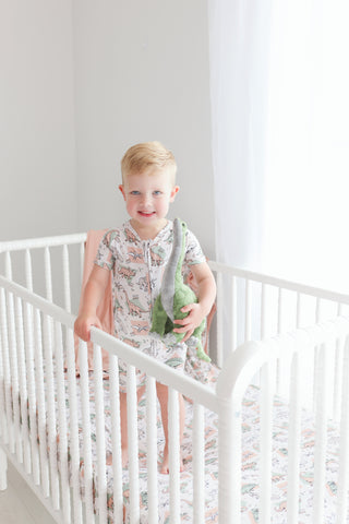 Toddler standing in white crib with multicolored dinosaur pattern on fitted sheet and pajamas.