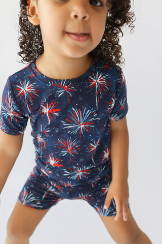 Child wearing navy short-sleeve shirt and matching shorts with red, white and blue firework print throughout.