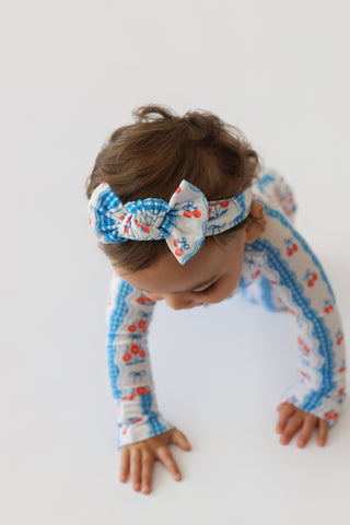 Toddler wearing white romper with blue gingham and cherry print sleeves and matching bow headband.