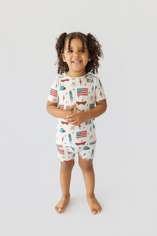 Child wearing cream short set with American flags, skateboards, and patriotic print throughout.