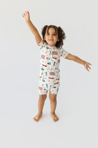 Toddler wearing cream short set with red, blue, and white patriotic print including flags, skateboards, and palm trees.