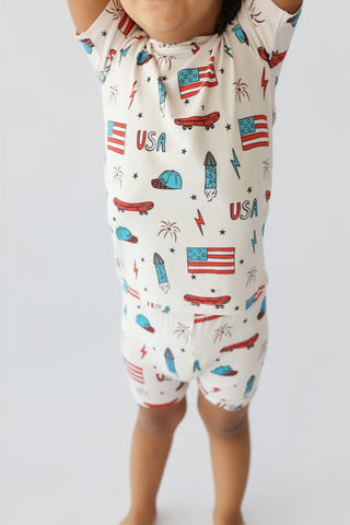 Child wearing cream short set with red and blue skateboard, flag, and star print throughout.