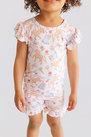 Child wearing white short set with ruffled sleeves and peach horses, boots, flowers, and bows print.