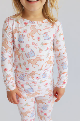 Child wearing long-sleeve pajama set with horses, boots, flowers, and carrots printed in orange and pink on white.