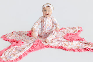 Infant sitting on a pink ruffle-edged blanket with floral and boot print pattern, wearing matching pajamas and headband.