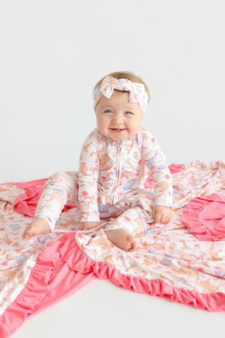 Baby wearing cream pajamas with boot and floral print, matching headband bow, sitting on pink ruffled blanket.